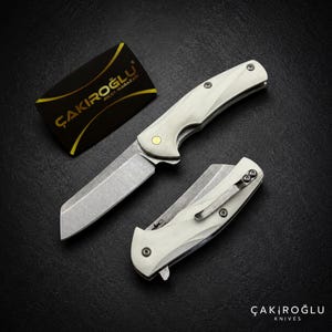 Könnte beinhalten: Zwei Klappmesser mit cremefarbenen Griffen und Metallklingen. Ein Messer ist geöffnet und zeigt eine gerade Klinge, während das andere geschlossen ist und einen Metallclip besitzt. Eine schwarze Karte mit dem goldenen Text ÇAKIROĞLU BIÇAK MAGAZASI.