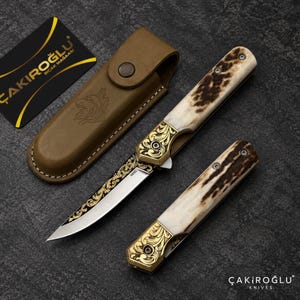 Könnte beinhalten: Ein Klappmesser mit Hirschhorngriff, Messingbacke mit komplexen Schnörkeln und einer Stahlklinge mit goldfarbenen Blumenverzierungen. Enthält eine braune Lederscheide mit Wolfskopf-Logo und eine schwarze Karte mit dem Text ÇAKIROĞLU BIÇAK MAĞAZASI.