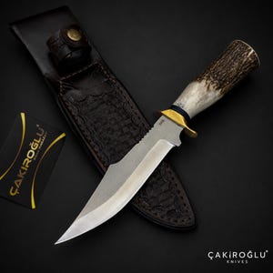 Puede incluir: Un cuchillo de hoja fija con mango de asta texturizada, guarda de latón y hoja de acero plateado. Se acompaña de una funda de cuero marrón oscuro con detalles en relieve y una tarjeta negra con el texto ÇAKIROĞLU BIÇAK MAĞAZASI.