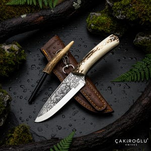 Puede incluir: Un cuchillo de hoja fija con mango de asta texturizada y hoja de acero plateado con intrincados patrones grabados y el texto N690. El cuchillo se acompaña de una funda de cuero marrón y un encendedor de fuego con mango de asta a juego.