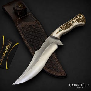 Peut inclure: Un couteau à lame fixe avec une lame en acier incurvée marquée N690 et un manche en bois de cerf texturé avec des goupilles vertes. Le couteau repose sur un étui en cuir marron foncé avec un motif à carreaux estampé et une fermeture à pression.