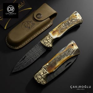Puede incluir: Dos cuchillos plegables con detalles dorados ornamentados y hojas estampadas se muestran con una funda de cuero marrón. Los mangos de los cuchillos tienen un aspecto natural y moteado. La marca "ÇAKIROĞLU KNIVES" es visible.
