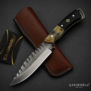 Peut inclure: Un couteau à lame fixe avec une lame en acier texturé, un manche bicolore en bois sombre et corne claire, et un étui en cuir marron gravé d'un loup. Une carte noire avec le texte ÇAKIROĞLU est visible à côté du couteau.