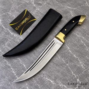 Könnte beinhalten: Ein feststehendes Messer mit einer silbernen Stahlklinge mit zwei parallelen Rillen, einem schwarzen Griff mit goldfarbenen Metallakzenten und einer schwarzen Scheide. Eine kleine schwarze Karte mit dem goldenen Text ÇAKIROĞLU BIÇAK MAĞAZASI liegt daneben.