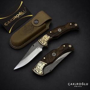 Può includere: Due coltelli a serramanico con manici in legno scuro, bolster in ottone con incisioni floreali e lame in acciaio testurizzato. Un coltello è aperto, l'altro è chiuso. Un fodero in pelle beige con un lupo impresso e una carta nera ÇAKIROĞLU.