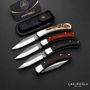 Peut inclure: Quatre couteaux pliants avec des lames en acier N690 et des manches variés en bois et en bois de cerf, accompagnés d'un étui en cuir marron foncé et d'une carte de visite noire portant le texte ÇAKIROĞLU BIÇAK MAĞAZASI.