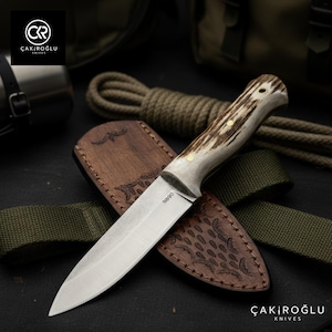 Puede incluir: Un cuchillo de caza con un mango texturizado de color claro y una hoja de acero inoxidable. El cuchillo descansa en una funda de cuero marrón con un diseño en relieve. La hoja está marcada con "N890". Producto de ÇAKIROĞLU KNIVES.
