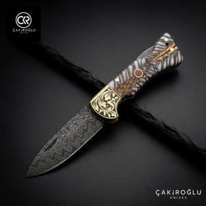 Könnte beinhalten: Ein Klappmesser mit gemusterter Klinge und dekorativem Griff. Der Griff hat goldfarbene Metallakzente und ein weißes und braunes Design. Das Messer ist vor einem dunklen Hintergrund platziert, mit dem Text "ÇAKIROĞLU KNIVES" sichtbar.