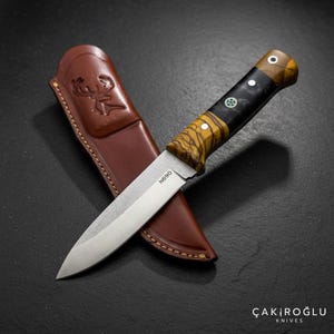 Puede incluir: Un cuchillo de caza con mango de madera y funda de cuero. La hoja del cuchillo es plateada y el mango tiene un patrón negro y amarillo. La funda es marrón y está grabada con un emblema de ciervo. El texto "N690" es visible en la hoja.