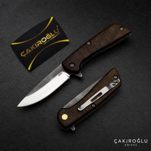 Könnte beinhalten: Ein Klappmesser mit dunklem Holzgriff und einer silbernen Klinge mit der Aufschrift N690, begleitet von einem geschlossenen Klappmesser mit silbernem Taschenclip und goldener Pivot-Schraube sowie einer schwarzen Karte mit dem Text ÇAKIROĞLU BIÇAK MAĞAZASI.