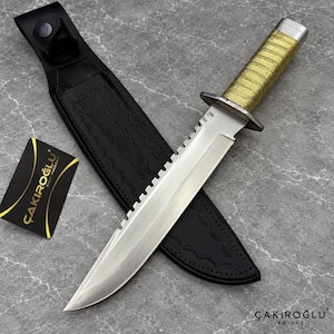 Könnte beinhalten: Ein feststehendes Messer mit einer silbernen Stahlklinge mit gezahntem Rücken, einem gold umwickelten Griff und einem Metallknauf. Es wird von einer schwarzen Lederscheide mit Ziernähten und einer schwarzen Karte mit der Aufschrift ÇAKIROĞLU begleitet.