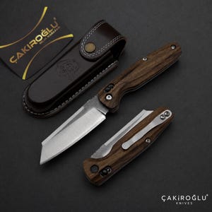 Puede incluir: Dos navajas plegables con mangos de madera oscura y hojas de metal plateado, una abierta y otra cerrada con un clip de bolsillo plateado. Se incluyen una funda de cuero marrón oscuro con botón a presión y una tarjeta negra con el texto ÇAKIROĞLU BIÇAK MAĞAZASI.