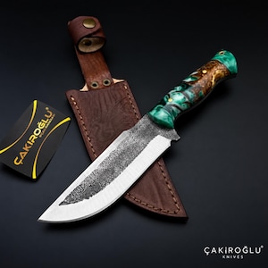 Peut inclure: Un couteau à lame fixe avec une finition métallique sombre et texturée sur la lame et un manche aux motifs de résine verte et brune tourbillonnants. Il est accompagné d'un étui en cuir brun et d'une carte noire avec le texte ÇAKIROĞLU.