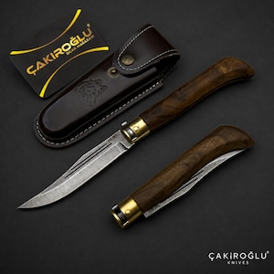 Può includere: Due coltelli a serramanico con manici in legno scuro e bolster in ottone, uno aperto e uno chiuso. Il coltello aperto ha una lama in acciaio damascato con la scritta M600. Un fodero in pelle marrone con emblema di lupo e una carta nera.