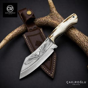 Puede incluir: Un cuchillo de acero inoxidable con mango de hueso y funda de cuero marrón. La hoja presenta un diseño de águila grabado. El cuchillo está etiquetado como "N690" y se muestra sobre una superficie oscura.