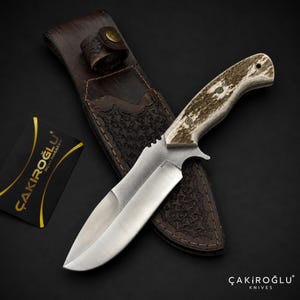Peut inclure: Un couteau à lame fixe avec un manche en bois de cerf texturé et une lame en acier marquée N690, accompagné d'un étui en cuir marron foncé avec des motifs gaufrés et une fermeture à pression en laiton, et une carte noire ÇAKIROĞLU BIÇAK MAĞAZASI.