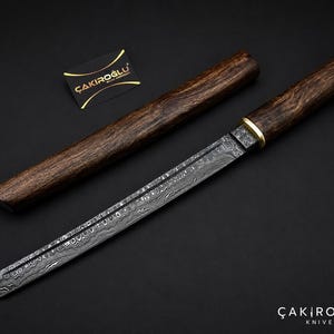 Può includere: Un coltello in acciaio Damasco con manico e fodero in legno. La lama presenta un motivo unico e il manico è marrone scuro con un accento dorato. Il fodero è anch'esso in legno marrone scuro. Un coltello fatto a mano.