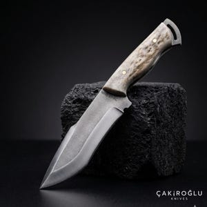 Puede incluir: Un cuchillo de caza artesanal con un mango texturizado de color claro y una hoja plateada. La hoja tiene un borde curvo y una punta. El texto "N690 60HRC" está grabado en la hoja. El cuchillo se exhibe sobre una superficie negra texturizada.