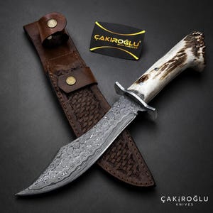 Puede incluir: Un cuchillo de caza con una hoja de acero estampada y un mango de hueso. El cuchillo descansa sobre una funda de cuero marrón con un patrón de tejido de cesta. También es visible una tarjeta de visita con el texto "ÇAKIROĞLU KNIVES".