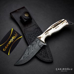 Puede incluir: Un cuchillo de hoja fija con un patrón de acero de Damasco oscuro y ondulado en la hoja y un mango de asta de color crema texturizado asegurado con tres pasadores de latón. Incluye una funda de cuero negro y una tarjeta negra con el texto ÇAKIROĞLU.