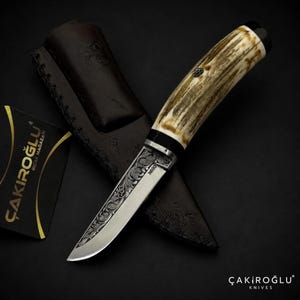Könnte beinhalten: Ein feststehendes Messer mit einem strukturierten, cremefarbenen Griff und einer silberfarbenen Klinge mit komplexen floralen Gravuren und dem Text N690. Es wird von einer dunkelbraunen Lederscheide und einer schwarzen Karte mit dem Text ÇAKIROĞLU BIÇAK MAĞAZASI begleitet.