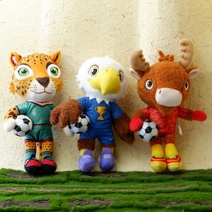 Könnte beinhalten: Drei Plüschtierfiguren: ein Jaguar, ein Adler und ein Elch, die jeweils einen Fußball halten. Der Jaguar ist in Grün und Gold, der Adler in Blau und der Elch in Rot gekleidet. Alle tragen Fußballuniformen und haben detaillierte Gesichtszüge.