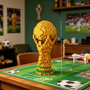 May include: A yellow, block-style trophy, 15cm (5.91 inches) tall and 6cm (2.36 inches) wide, sits on a green and white soccer field-themed mat. The trophy is a detailed replica of the World Cup trophy. The background includes soccer-themed decor.