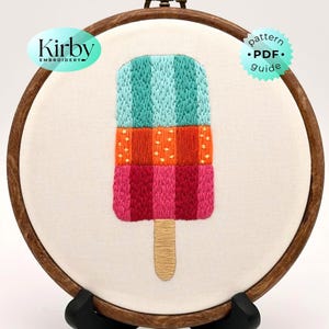 May include: A circular wooden embroidery hoop containing white fabric with a hand-stitched popsicle design. The popsicle features vertical stripes in teal, orange with yellow dots, and magenta, with a tan wooden stick. Text reads Kirby Embroidery.