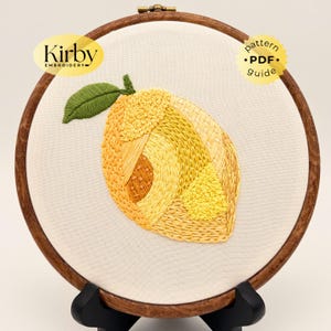 May include: Embroidered lemon design on a white fabric stretched within a wooden embroidery hoop. The lemon is yellow and orange with a green leaf. The text "Kirby Embroidery" and "pattern PDF guide" are also visible.