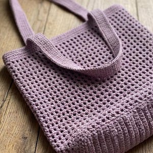 May include: A lavender-colored, crocheted tote bag with a rectangular body and two sturdy handles. The bag features a mesh-like pattern throughout, with a solid, ribbed base and top edge. The handles are also crocheted in a matching lavender color.