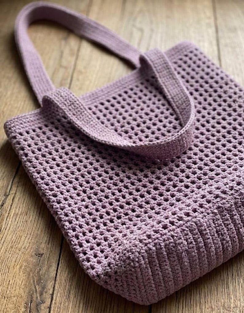 May include: A lavender-colored, crocheted tote bag with a rectangular body and two sturdy handles. The bag features a mesh-like pattern throughout, with a solid, ribbed base and top edge. The handles are also crocheted in a matching lavender color.