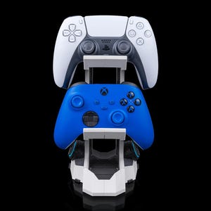 Könnte beinhalten: Ein weiß-schwarzer PlayStation 5-Controller und ein blauer Xbox-Controller auf einem weiß-schwarzen Ladeständer. Die Controller befinden sich vor einem schwarzen Hintergrund.