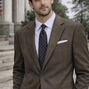 May include: A brown plaid suit jacket with a white pocket square, paired with a blue dress shirt and a patterned navy tie. The suit is worn with light-colored pants. The man is wearing a silver watch.