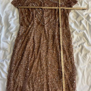 Puede incluir: Un vestido de lentejuelas en color oro rosa con mangas cortas y cuello redondo, que presenta un intrincado trabajo de cuentas con un patr&oacute;n geom&eacute;trico en el corpi&ntilde;o. Una regla de madera mide 56 cm de ancho sobre el pecho y el largo.