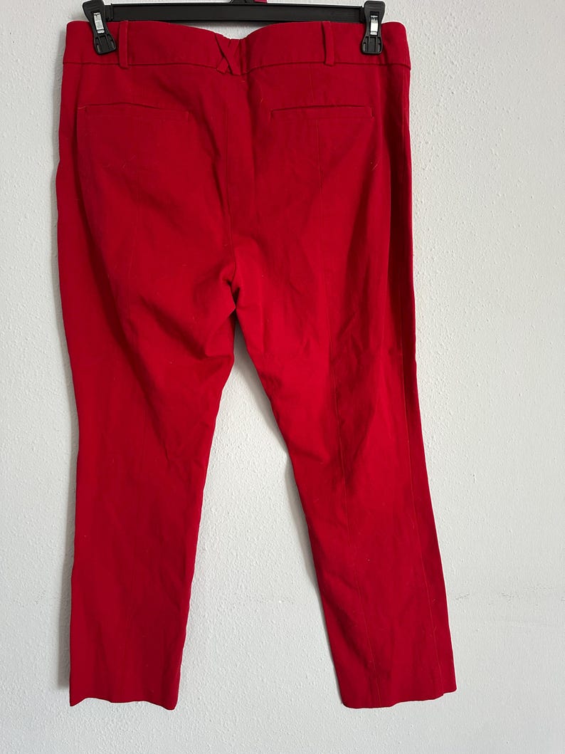May include: Bright red, tailored pants hanging on a black hanger. The pants have a classic design with a straight leg and a back pocket. The fabric appears to be a smooth, woven material.