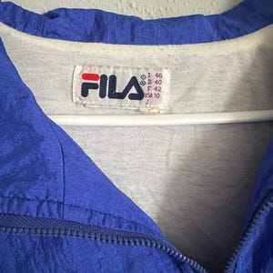 May include: A close-up of a blue Fila jacket with a white interior. The jacket features a zipper and a Fila logo patch. The tag shows size details: I 46, D 40, F 42, USA 10, and J.