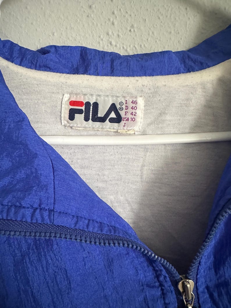 May include: A close-up of a blue Fila jacket with a white interior. The jacket features a zipper and a Fila logo patch. The tag shows size details: I 46, D 40, F 42, USA 10, and J.