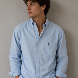 May include: Light blue button-down shirt with long sleeves. The shirt features a small pocket on the chest and a dark blue embroidered logo. The shirt is made of a soft, light fabric.
