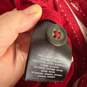 May include: Close-up of red fabric with a black tag and a button. The tag lists the fabric composition: 52% cotton, 41% rayon, and 7% spandex. The button is dark brown and attached with red thread.