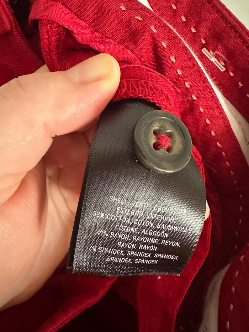 May include: Close-up of red fabric with a black tag and a button. The tag lists the fabric composition: 52% cotton, 41% rayon, and 7% spandex. The button is dark brown and attached with red thread.