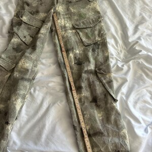 May include: Cargo pants featuring a mottled gray and green camouflage-style pattern, multiple flap pockets on the thighs and sides, and a button fly. A wooden ruler is placed across the waist and down the leg to show the garment's dimensions.