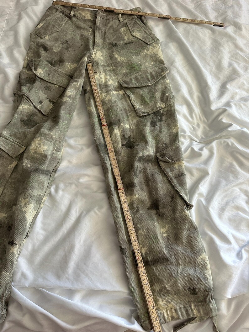 May include: Cargo pants featuring a mottled gray and green camouflage-style pattern, multiple flap pockets on the thighs and sides, and a button fly. A wooden ruler is placed across the waist and down the leg to show the garment's dimensions.