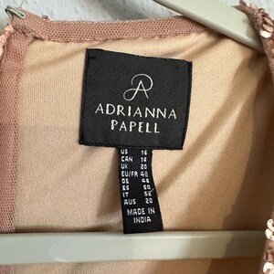 Puede incluir: Una prenda de color rosa empolvado con lentejuelas y una etiqueta de tela negra con el texto Adrianna Papell. La etiqueta muestra informaci&oacute;n de tallas como US 16, CAN 16, UK 20, EU/FR 48, DE 46, ES 50, IT 52, AUS 20 y Made in India.