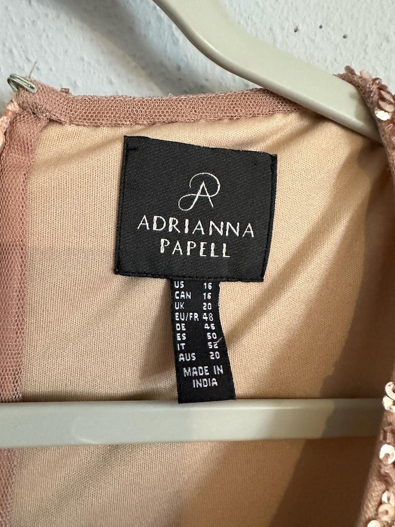 Puede incluir: Una prenda de color rosa empolvado con lentejuelas y una etiqueta de tela negra con el texto Adrianna Papell. La etiqueta muestra informaci&oacute;n de tallas como US 16, CAN 16, UK 20, EU/FR 48, DE 46, ES 50, IT 52, AUS 20 y Made in India.