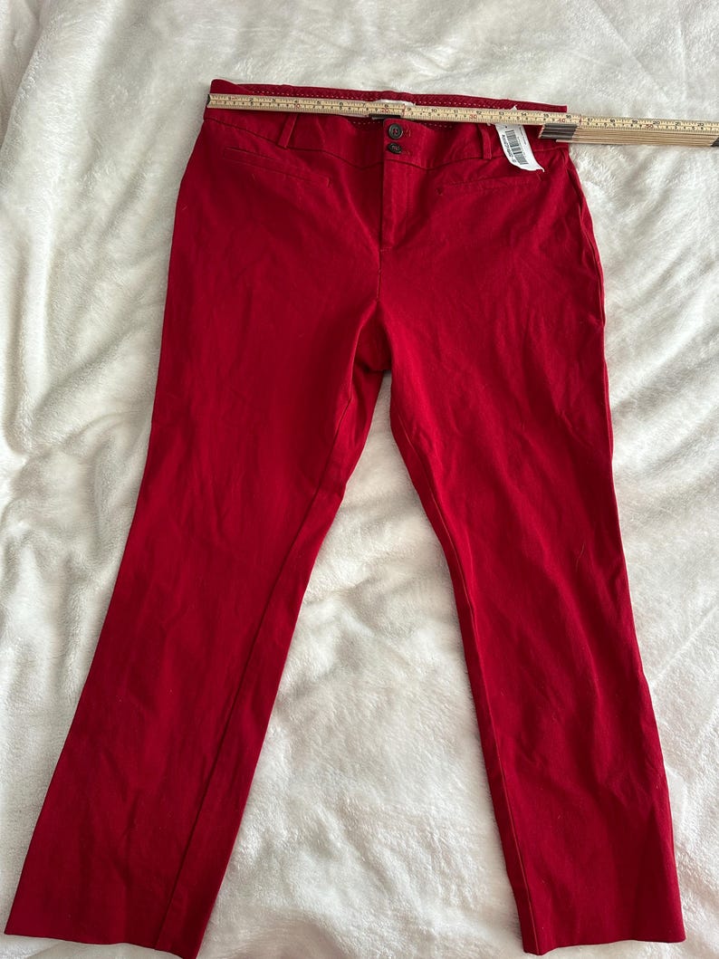 May include: Bright red pants with a button closure and a zipper. The pants are made of a solid fabric and have a classic, tailored design. A measuring tape is placed across the top of the pants.