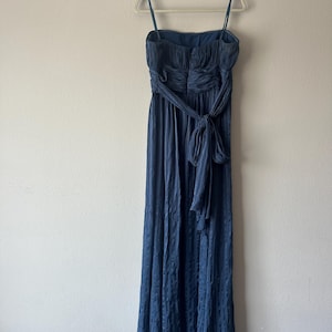 Peut inclure: Une robe de soir&eacute;e bleu marine avec des bretelles spaghetti et une taille cintr&eacute;e. La robe pr&eacute;sente une jupe fluide et une ceinture assortie nou&eacute;e en n&oelig;ud. La robe est suspendue &agrave; un cintre blanc contre un mur uni.
