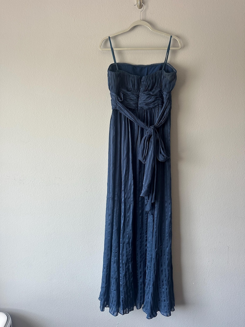 Peut inclure: Une robe de soir&eacute;e bleu marine avec des bretelles spaghetti et une taille cintr&eacute;e. La robe pr&eacute;sente une jupe fluide et une ceinture assortie nou&eacute;e en n&oelig;ud. La robe est suspendue &agrave; un cintre blanc contre un mur uni.