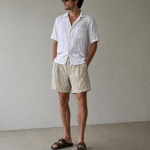 May include: A person wearing a white short-sleeved shirt, beige shorts, and brown sandals. The shirt has a single pocket on the chest. The shorts are pleated. The sandals have two straps.