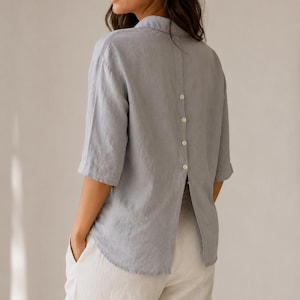 May include: A light gray button-down shirt with elbow-length sleeves and a vertical row of four small white buttons running down the center of the back, featuring a split hem design that opens from the bottom button to the lower edge of the shirt.