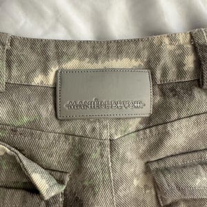 May include: A pair of grey denim pants featuring a mottled, tie-dye style pattern with subtle hints of light green. A rectangular grey leather-like patch is stitched to the back waistband, embossed with the text MANIERE DE VOIR in capital letters.